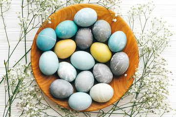 Stylish Easter eggs with spring flowers in wooden plate flat lay on white wooden background. Modern colorful easter eggs painted with pastel natural dye. Happy Easter, greeting card