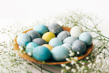 Fototapeta na wymiar Stylish Easter eggs with spring flowers in wooden plate on white wooden background. Modern easter eggs painted with natural dye in yellow,blue,green,grey marble colors. Happy Easter greetings