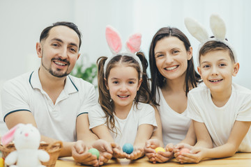 Inspired family in white t-shirts sitting together at the table, holding colored easter eggs in their hands, looking excitedly at the camera .