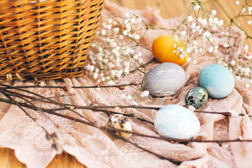 Stylish Easter eggs with spring flowers and willow branches on rustic fabric at wicker basket with holiday food in sunny light. Modern easter eggs painted with natural dye. Happy Easter