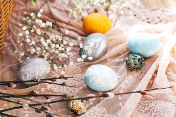 Fototapeta na wymiar Stylish Easter eggs with spring flowers and willow branches on rustic fabric at wicker basket with holiday food in sunny light. Modern easter eggs painted with natural dye. Happy Easter