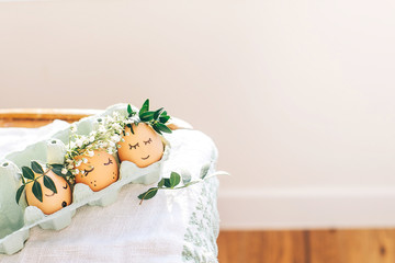 Happy Easter,  eco concept. Stylish Easter eggs with cute faces in floral wreath crowns in carton tray on rustic background. Modern easter eggs with flowers and sleepy eyes in sunny light