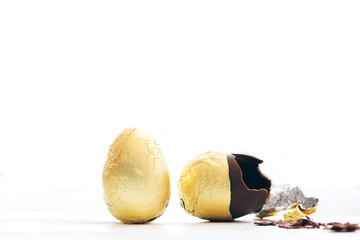 Stylish Easter egg in golden foil and broken chocolate egg with chocolate pieces in white light with space for text. Modern chocolate gold easter eggs. Happy Easter. Holiday gift.