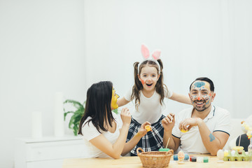 Cute little daughter with pigtails and bunny ears is hugging her parents. Mother and father painting easter eggs, playing around with paintbrushes and paint, colouring each others faces.