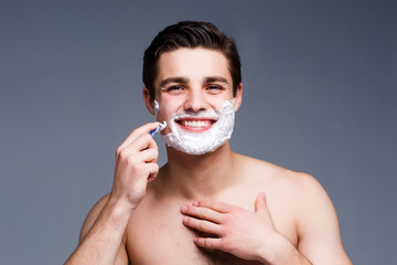 Young, attractive  man shaving .He is passing the razor for the beard while staying on grey background.