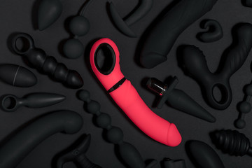 Erotic pleasure toys on a black background. Sex gadgets and masturbation devices. Pink dildo surrounded by black rubber and silicone sex toys: anal butt plugs, vibrators, vaginal balls. Top view.