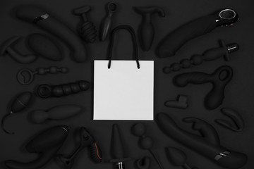 Shopping paper bag surrounded by black sex toys on a black background. Erotic pleasure toy. Sex gadget and masturbation device. Top view. Place for text. Flat lay. Sex shop concept.
