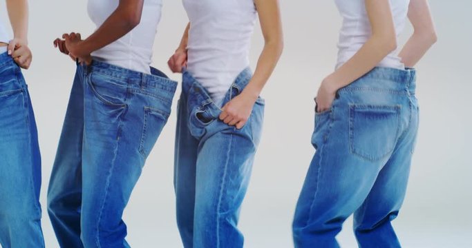 Slow motion of beautiful young smiling women of different ethnicities with perfect firm and slim body in white tank shirts dancing, having fun and showing weight loss by wearing old jeans. 