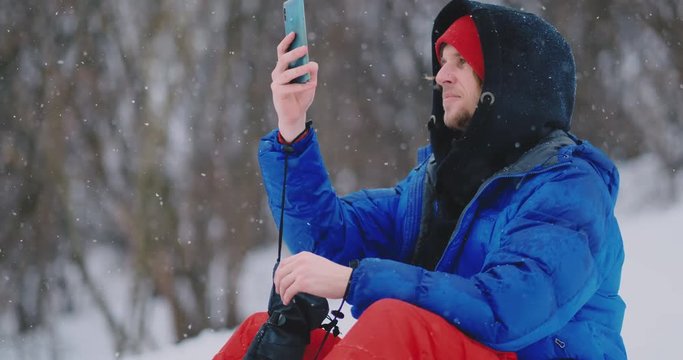 Male snowboarder sitting on the snow taking photos on the phone beautiful scenery of the resort for social networks