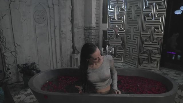 The beautiful young girl with long curly hair flaunts and shows off in bathtub filled with petals of roses, flirts, smiles, sultry look, happy, pleased with herself, makeup, sexy lips, wet hair