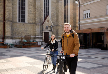 two people - friends or couple, pushing their bicycles in a old city square in Europe, Serbia, Novi Sad. Smiling and looking to camera.