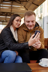 two friends, using one smartphone while holding it in a hand, in a cafe.