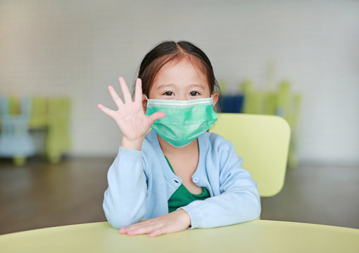 Little Asian Child Girl Wearing A Protective Mask With Showing Five Fingers Sitting On Kid Chair In Children Room.