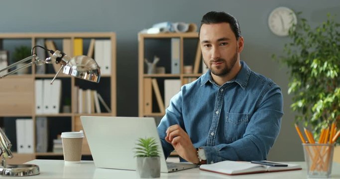 Portrait shot of the good looking stylish young Caucasian man working at the laptop computer in the office,then rising his head and smiling to the camera.