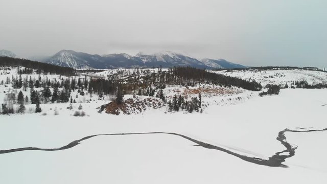 Drone flying above a frozen lake in Dillon Colorado. Flowing creeks cutting through the ice. Mountains in background. Cloudy dark dreary day. 1080p 30 fps.