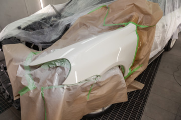 Painting the car body parts of the driver's white fender in the spray booth in the body repair shop, other elements are covered with film and paper from splashing.