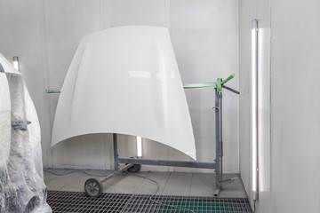 Repair of the big hood of a white car after an accident with the help of putty mounted on a special holder in a workshop for painting vehicles in a special chamber. Spray booth with bright lighting.