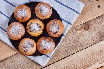 Cupcakes with chocolate chips with powdered sugar lie in a black baking sheet, on a napkin on a wooden background, flat lay