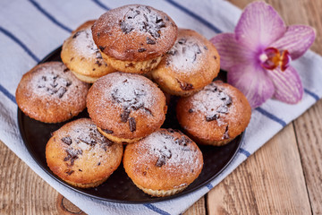 Cupcakes with chocolate chips with powdered sugar lie in a black baking sheet, on a napkin next to the orchid flower, on a wooden background. General view