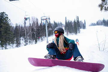 Woman snowboarder with dreadlocks it sitting with a snowboard on the ski slopes.