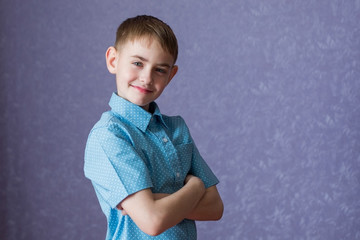 A boy in a blue shirt on a purple background is standing and folded two arms across his chest, smiling.