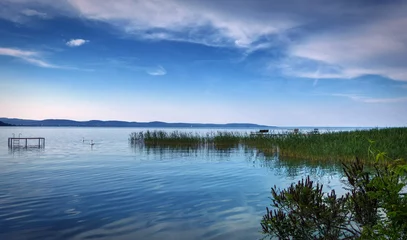 Foto op Canvas A tranquil scene at the Balaton lake, Hungary. Calm water, some green water grass, a few benches in the water, two swans, blue sky with some clouds, water reflections. © Photo Shots