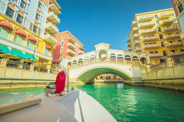 Fototapeta na wymiar Venetian bridge on canals of picturesque Qanat Quartier icon of Doha, Qatar from a touristic boat with flag of Qatar. Venice at the Pearl, Persian Gulf, Middle East. Famous attraction at sunset light.