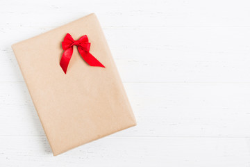 Present box in craft paper with red bow over white wooden background with copy space