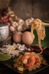 Thai fried noodles or pad thai with shrimp on black plate placed on the wood table there are eggs, garlic, bean sprouts, shallot, silver water bowl, chilli and cutting board placed back side.