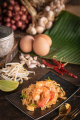 Thai fried noodles or pad thai with shrimp on black plate placed on the wood table there are eggs, garlic, bean sprouts, shallot, silver water bowl, chilli, fork, spoon and cutting board placed around