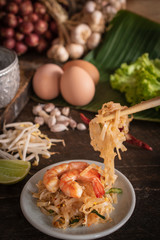 Thai fried noodles or pad thai with shrimp on white plate placed on the wood table there are eggs, garlic, bean sprouts, shallot, silver water bowl, chilli and cutting board placed back side. 