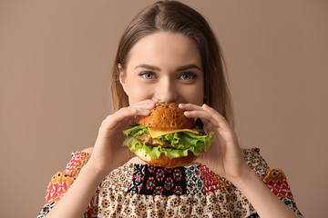 Beautiful young woman eating tasty burger on color  background