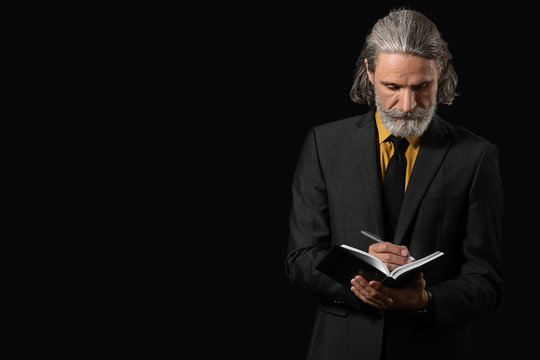 Handsome mature businessman writing in notebook on dark background