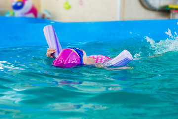 A female child in a pink rubber hat and blue glasses swims in the back pool with an arc for swimming.