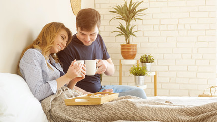 Young couple has breakfast in bed drinks tasty coffee miling in bedroom, sunlight