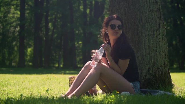 Young woman with wireless headphones listens to music and drinks mineral water from plastic bottle. Girl student sitting on grass in city park among trees and foliage on a bright sunny day.
