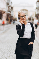 Beautiful trendy blond business woman in glasses with smartphone and handbag of documents in her hands outdoors.