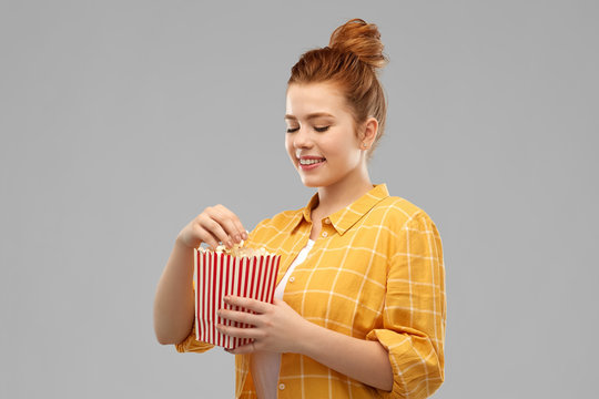 fast food and people concept - smiling red haired teenage girl in checkered shirt eating popcorn from striped bucket over grey background