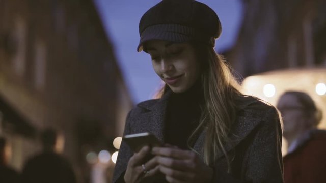 portrait of young beautiful fashionable brunette woman standing on the street at evening and using phone. Model wearing stylish gray coat, hat and black golf. HD stock footage.