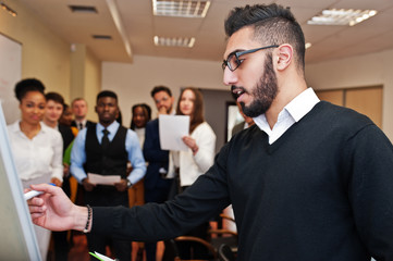 Arabian business coach presenting report standing near whiteboard pointing on sales statistic shown on diagram and chart teach diverse multiracial company members gathered together in conference room.