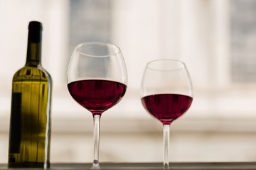 Two glasses of red wine and a bottle on a table next to a window