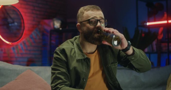 Portrait shot of the happy Caucasian young man in glasses watching sport channel on TV in the evening, drinking beer and smiling.