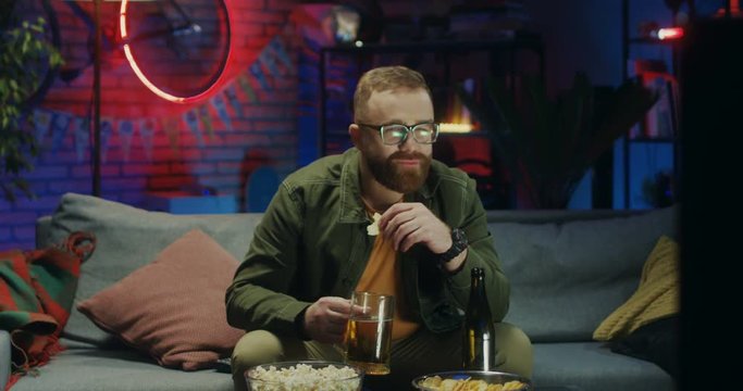 Caucasian young guy in glasses sitting on the couch with beer and snacks an worrying about sport match on TV at night.