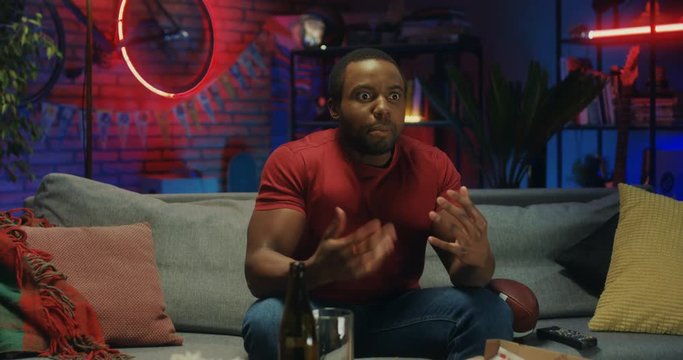 African American young cheerful guy sitting on the sofa in the dark cozy living room and watching sport game on TV, cheering up when his team winning or scoring a goal.