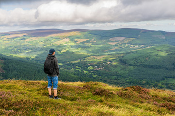 Naklejka na ściany i meble Tourist enjoying the beautiful view and resting on the top of Maulin Peak, Wicklow Mountains, after a hike on a cloudy summer day. 40 shades of green of Ireland.
