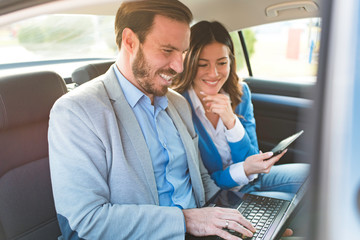 Two young business people driving on a backseat of a car working on laptop and smiling. Successful team businessman and businesswoman. 
