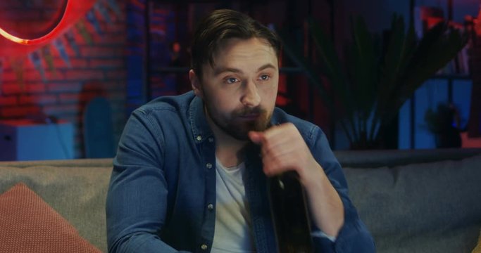 Portrait shot of the Caucasian good looking young caucasian guy sipping beer from a bottle and watching TV while changing channels in the dark living room in the evening.