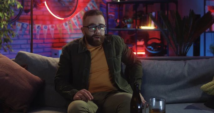 Caucasian young guy in glasses sitting on the couch in the dark room at night and watching scary movie on TV.
