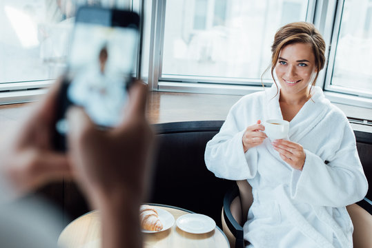 cropped view of man taking photo of attractive girlfriend holding cup while sitting in bathrobe in hotel