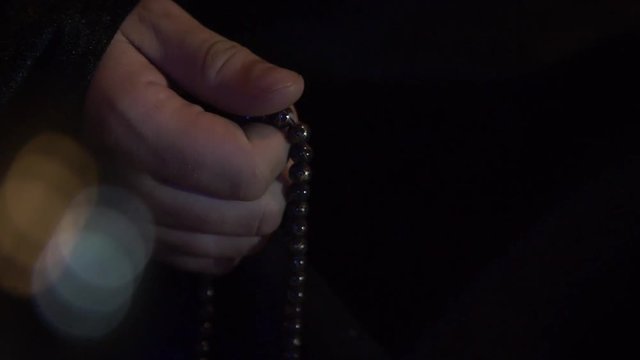 A man in a coat in a dark room turns the beads in his hand. Black beads with a small pattern. On the camera a few highlights of yellow and blue. Close-up. Slow motion.